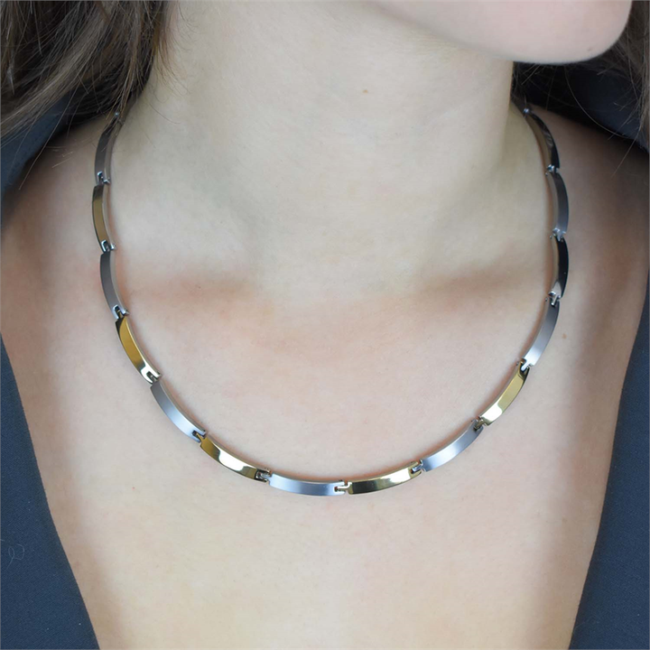 Een close-up van een persoon die de Boccia Titanium Collier Bicolor 0816-03 draagt, met afwisselend zilveren en gouden rechthoekige titanium schakels, tegen een achtergrond van blote huid en een donker, openvallend bovenstuk.
