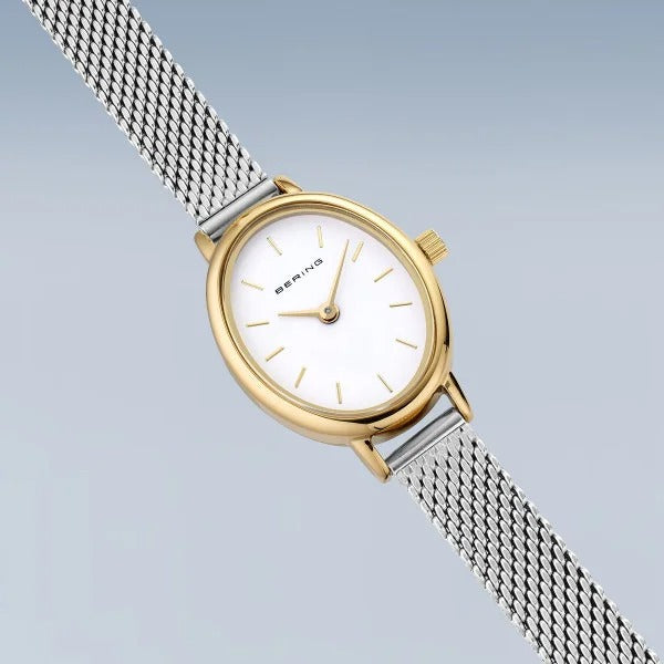 De Bering 11020-010 dameshorloge heeft een gouden en zilveren ontwerp, ovale witte wijzerplaat met gouden markeringen, saffierglas, quartz uurwerk en een zilveren mesh band, hier afgebeeld tegen een lichtblauwe achtergrond.