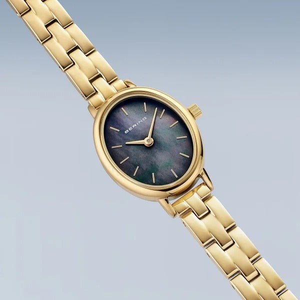 Het Bering dameshorloge 11020-732 van goudkleurig roestvrij staal heeft een ovale parelmoer-wijzerplaat, minimalistische gouden uurmarkeringen en een elegante schakelband, gepresenteerd tegen een lichtblauwe achtergrond.