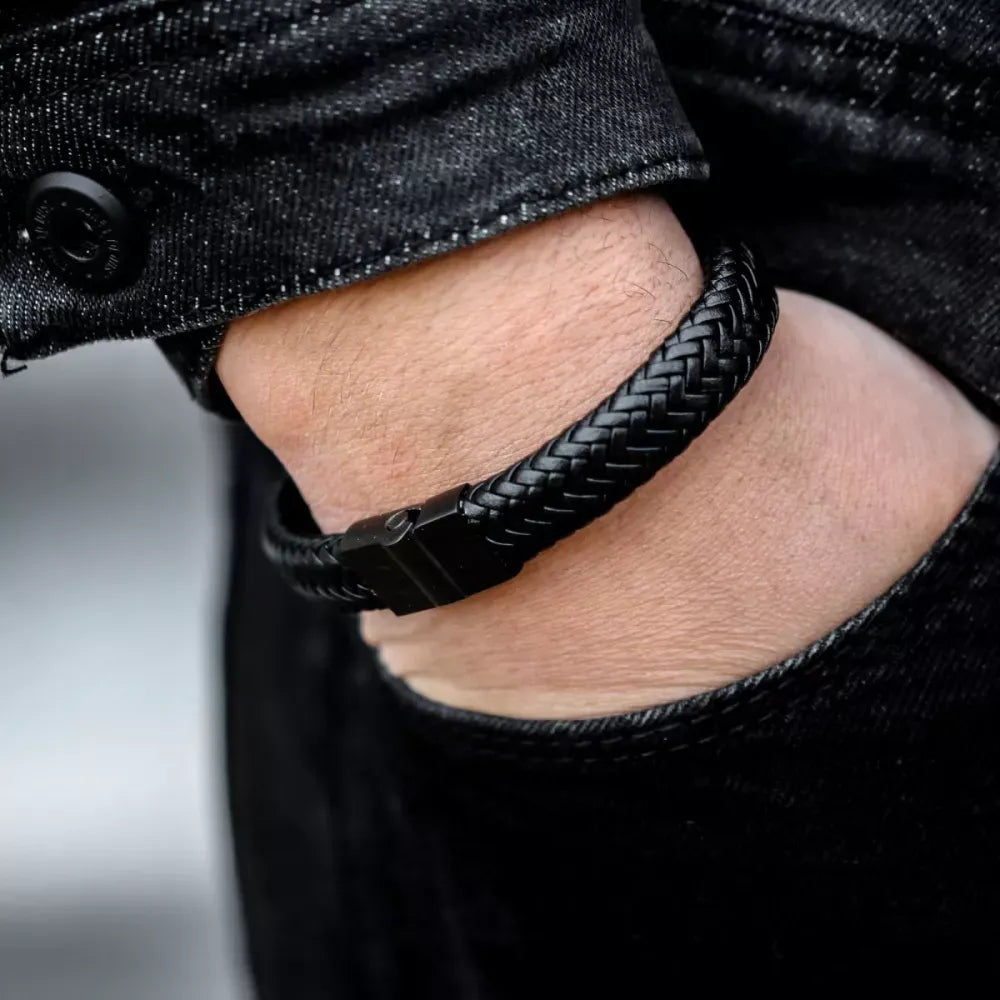 Iemand draagt de Rebel & Rose Full Metal Herringbone Black Black armband - een zwart, gevlochten stuk leer en roestvrij staal met een zwarte sluiting - in combinatie met een zwarte jeans en een zwart spijkerjack.