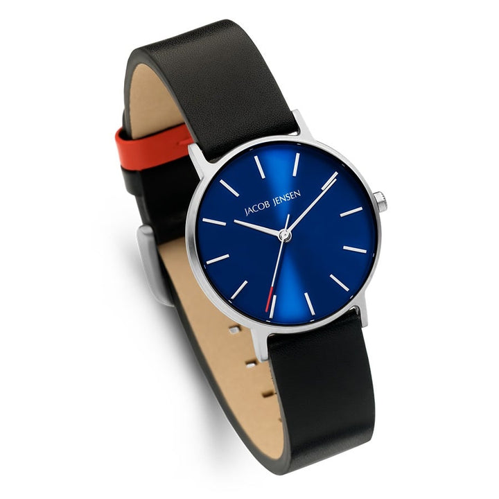 De Jacob Jensen 171 is een modern quartz polshorloge met een blauwe wijzerplaat met minimalistische witte uurmarkeringen en een klein rood accent op de secondewijzer. Het heeft een zwarte leren band met een rode onderkant en is omhuld met strak titanium voor stijl en duurzaamheid.