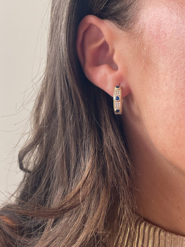 Een close-up van een vrouw met Excellent Jewelry's geelgouden collier met blauwe saffier (0.50 crt) en diamanten (0.24 crt), 14 karaat, met gouden hoepel oorbellen en bruin haar gedeeltelijk zichtbaar op de achtergrond.
