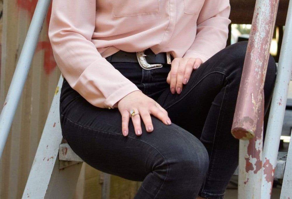 Een persoon in een lichtroze blouse, zwarte jeans en een western-stijl riem zit op een metalen trap. Hun handen rusten op hun schoot, waardoor de Ring vierkant en kubusje Goud|Geel van CLIC Creations wordt benadrukt. De achtergrond toont een deel van een leuning en structuur.