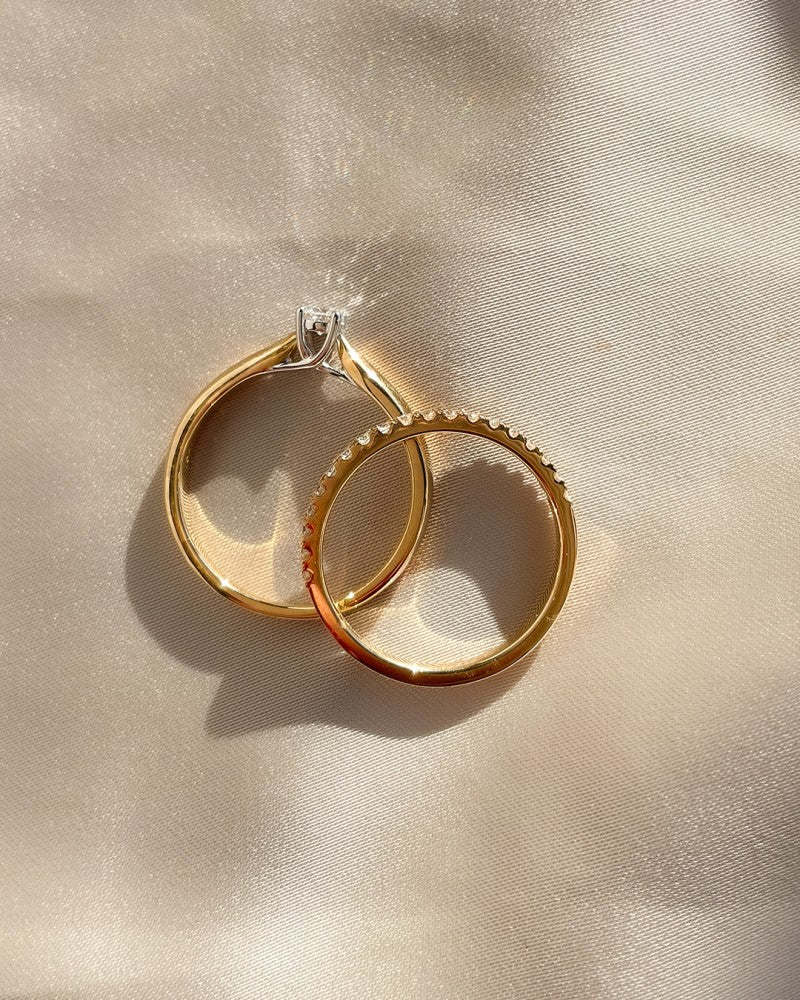Twee 14K geel- en witgouden ringen van Excellent Jewelry rusten op glad satijn. De ene is voorzien van een 0,15 ct briljante diamant; de andere heeft een gestructureerde rand. Beide werpen zachte schaduwen over de lichtgekleurde stof onder warme verlichting.