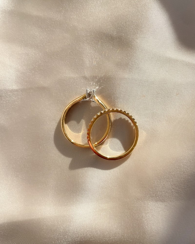 Twee gouden ringen van Excellent Jewelry rusten op zachte stof in het zonlicht: een effen en de Elegante Bicolor Gouden Ring met Briljanten 0.19 crt., voorzien van een subtiele getextureerde rand voor een elegante touch.