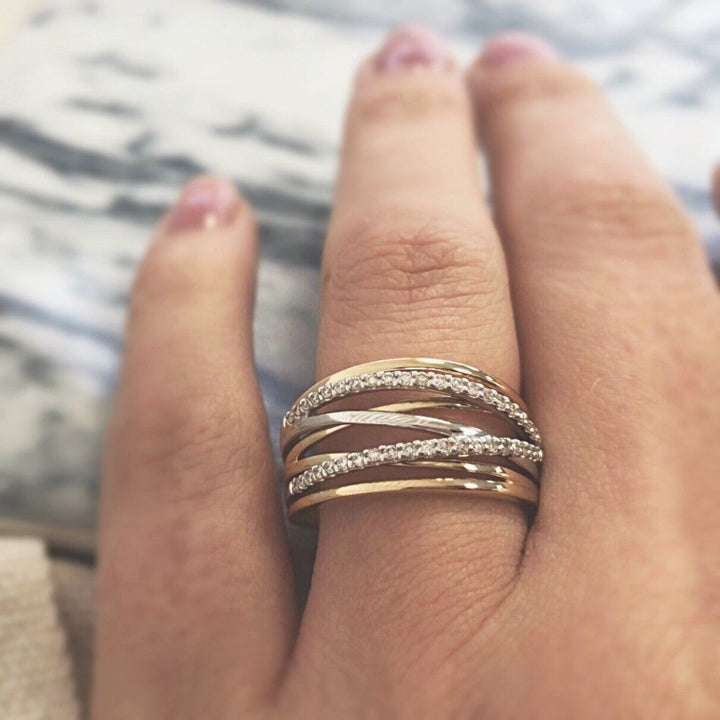 Een hand met roze nagellak showt de Excellent Jewelry Bicolor Gouden Crossover Ring met Diamant - 14 karaat, met kriskras door elkaar lopende banden, sommige bezet met diamanten, tegen een onscherpe achtergrond.