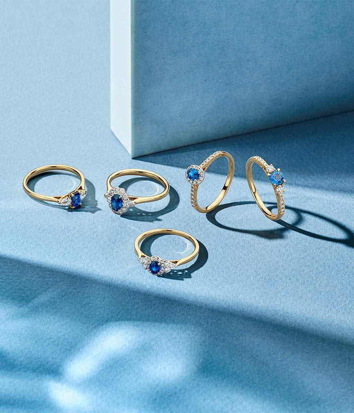 Vijf 14-Karaats Bicolor Gouden Damesringen met Blauwe Saffier en Vintage Diamanten Zetting van Excellent Jewelry staan afgebeeld op een lichtblauw oppervlak met schaduwen; sommige staan rechtop, andere liggen plat, met een wit geometrisch blok op de achtergrond.