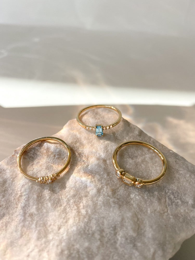 Drie delicate gouden ringen, waaronder de Excellent Jewelry "Ring geelgoud briljant 0.10 crt." en een met een rechthoekige blauwe steen, zijn gerangschikt op een gestructureerd beige stenen oppervlak in zacht natuurlijk licht.
