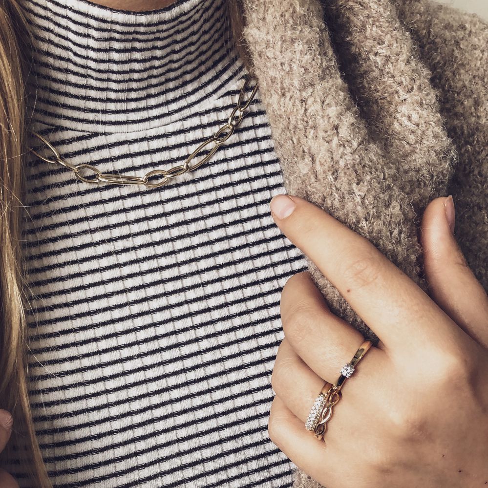 Een persoon draagt een gestreepte coltrui, een dikke gouden ketting en de Excellent Jewelry Stijlvolle Geelgouden Ring met Briljant in Pavézetting - 14 Karaat Goud, 0.32ct Diamant, met een beige trui gedrapeerd over één schouder.