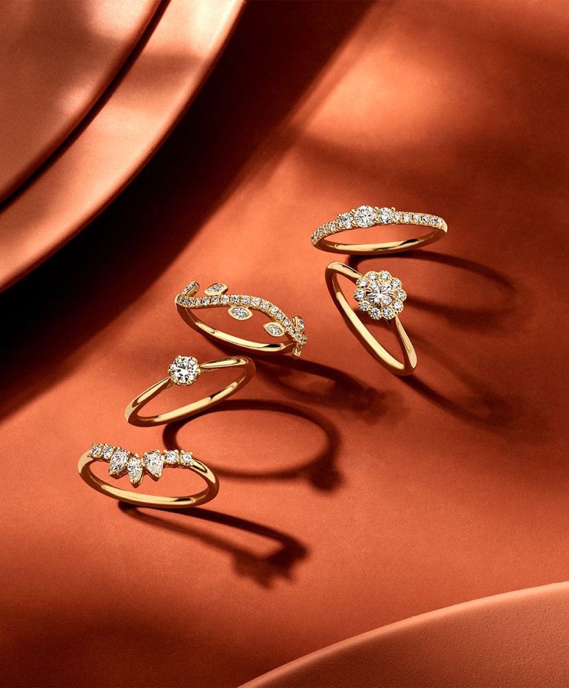 De Geelgouden Driesteens Ring met Extra Diamanten in de Scheen voor Maximale Schittering van Excellent Jewelry schittert op een koperen ondergrond, waarbij diamanten het geelgoud prachtig accentueren.