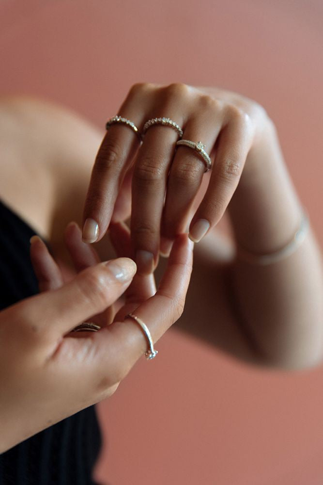Een close-up van gemanicuurde handen die de Witgouden Ring met Diamanten dragen: Subtiele Elegantie in 14 Karaat van Excellent Jewelry, met delicate zilveren ringen en een onscherpe roze achtergrond. De ene hand raakt de andere zachtjes aan.