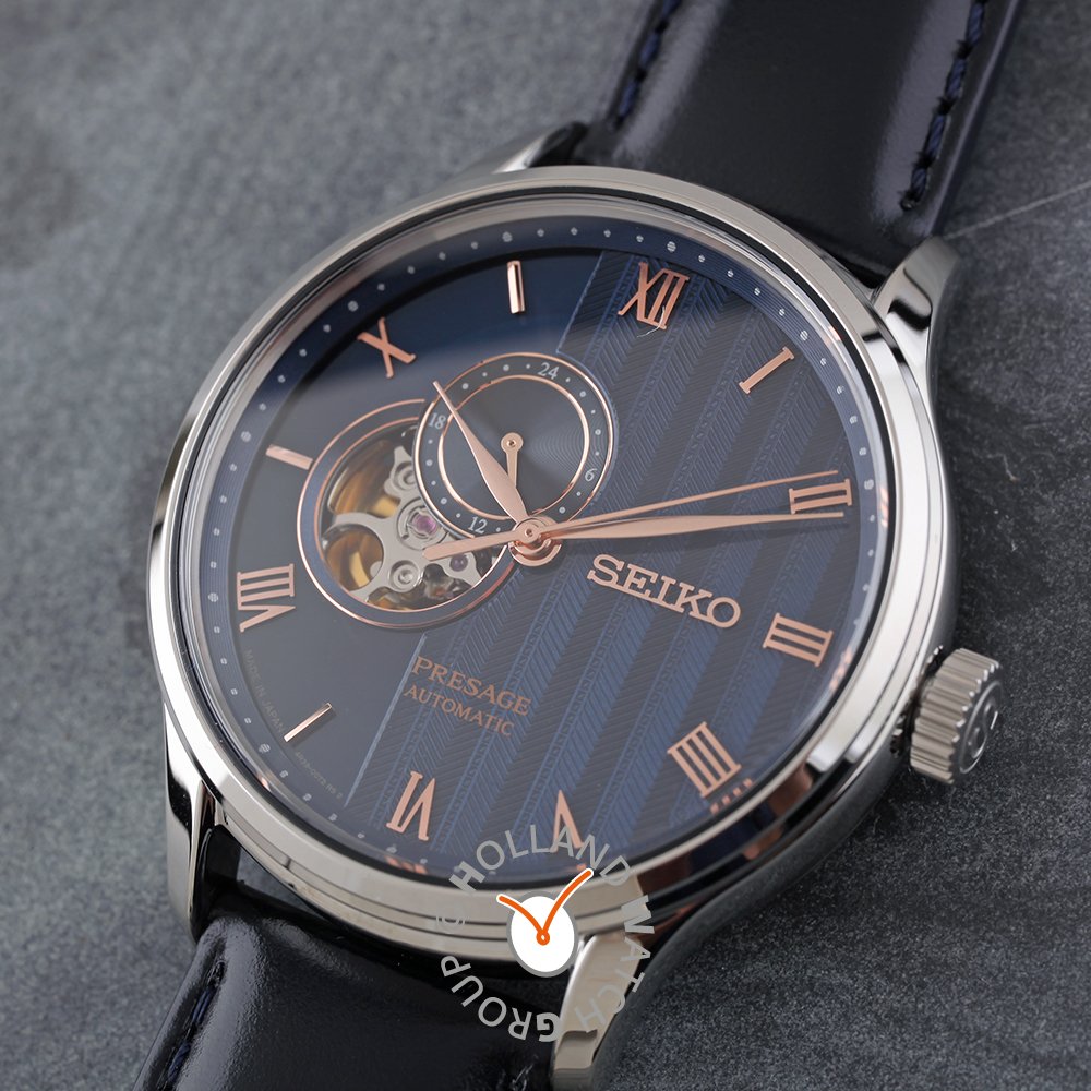 Een close-up van de Seiko heren horloge SSA421J1 uit de Presage lijn, met een automatisch uurwerk, blauw gedessineerde wijzerplaat met roségouden Romeinse cijfers, open-hart detail en een zwarte leren band op een grijs gestructureerd oppervlak.