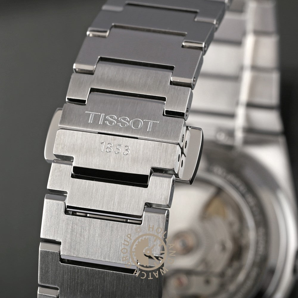 Close-up van een roestvrijstalen Tissot PRX Powermatic 80 horlogeband met "TISSOT 1853" gegraveerd op de sluiting; op de onscherpe achtergrond zijn een deel van de open achterkant van de horlogekast en het Zwitserse automatische uurwerk zichtbaar.
