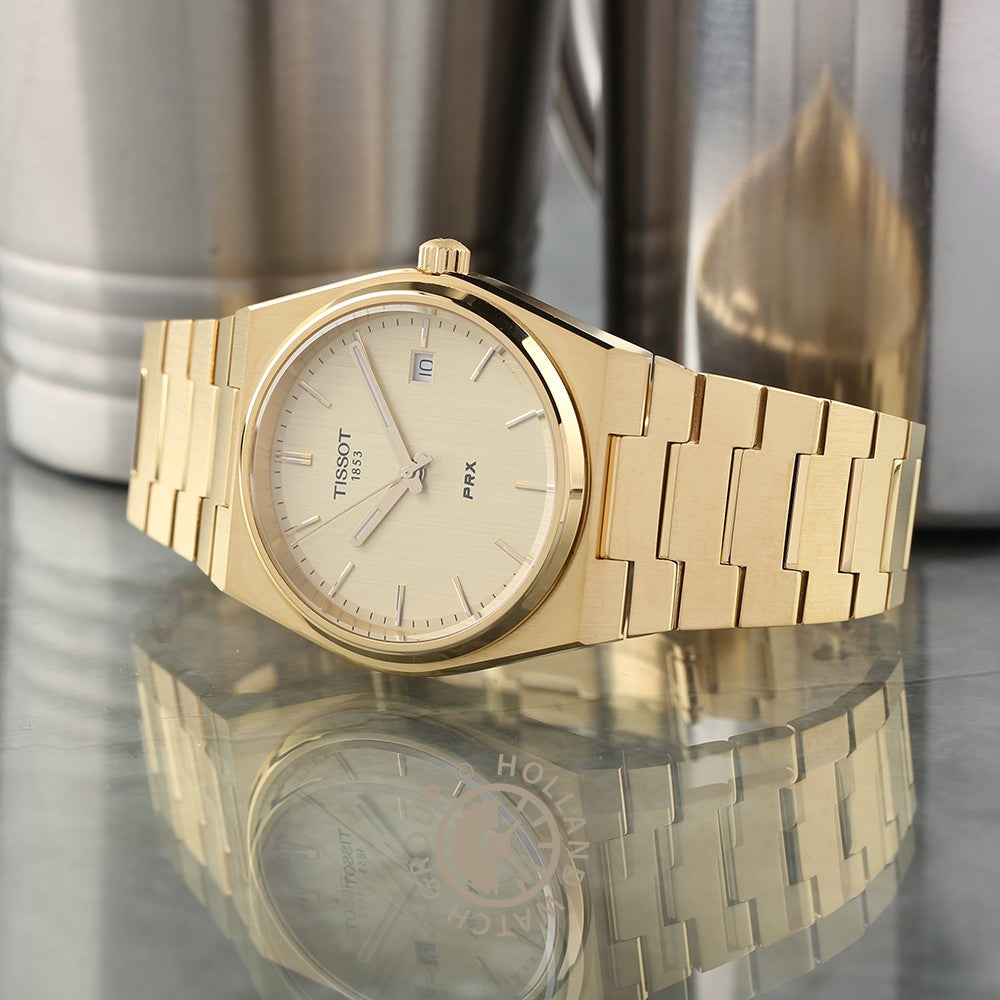 Een gouden Tissot PRX polshorloge van Tissot met Saffierglas, een champagnekleurige wijzerplaat en datumweergave rust op een reflecterend oppervlak, metalen cilindervormige vormen vervaagd op de achtergrond.