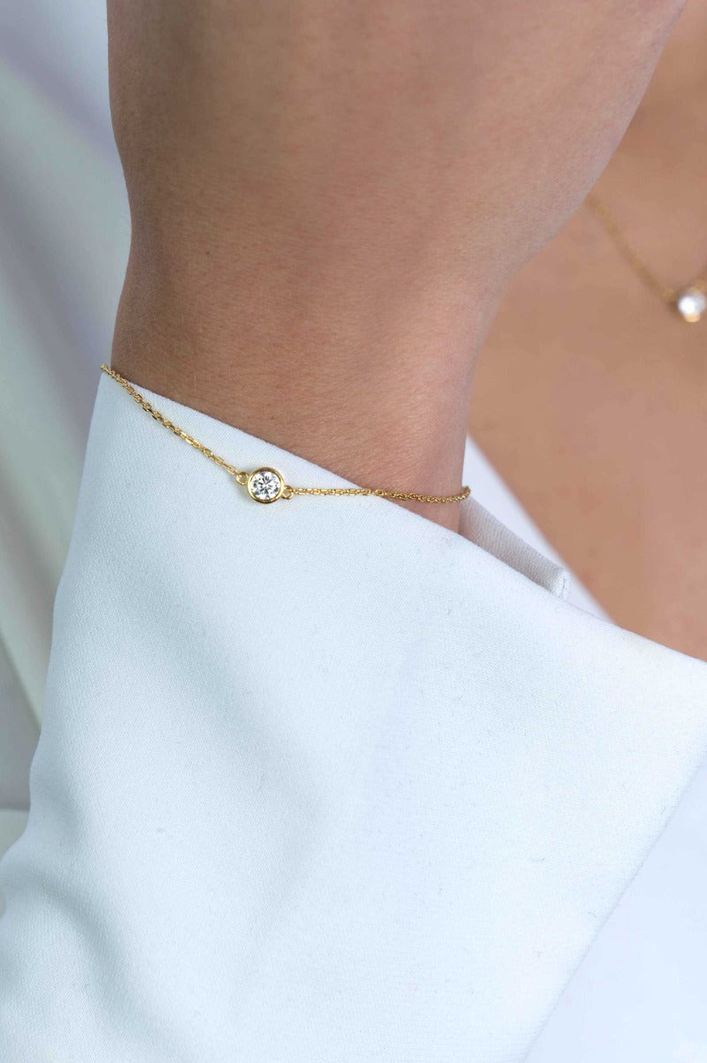 Close-up van een persoon die de ZINZI Armband Gold Plated Zia1393 Y draagt, met een delicaat gouden ontwerp met een kleine ronde diamant in een rond frame, elegant rustend om de pols over een witte mouw tegen een zacht wazige achtergrond.