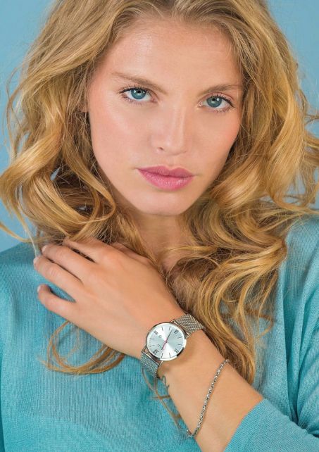 Een vrouw met golvend blond haar draagt een lichtblauw topje en een Zinzi Zilverkleurig Horloge Staal Ziw502 M, waarbij ze zachtjes haar sleutelbeen aanraakt terwijl ze naar de camera kijkt tegen een bijpassende lichtblauwe achtergrond.